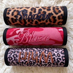 Op de afbeelding: Drie gepersonaliseerde handvatwraps. De bovenste wrap heeft een bruin-zwarte luipaardprint met de naam "Shadow" in blauw geborduurd. De middelste wrap is roze camouflage met "Blaze" in roze geborduurd. De onderste wrap is roze luipaardprint met "Lily M" in bruin geborduurd.