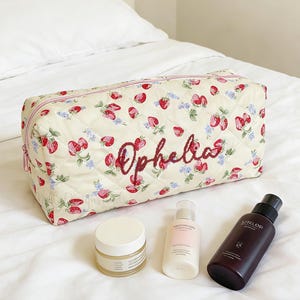 May include: Cream-colored quilted cosmetic bag with a strawberry and floral pattern, the name "Ophelia" embroidered in red. The bag is next to a jar, a bottle, and a spray bottle.