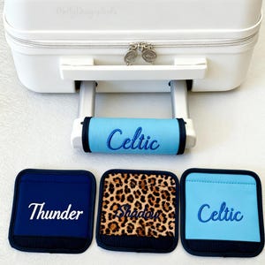 Op de afbeelding: Een witte reiskoffer met een uitschuifbare handgreep en drie handgreepbeschermers. Eén handgreepbeschermer is lichtblauw met "Celtic" in blauw geborduurd. Twee vierkante handgreepbeschermers zijn marineblauw met "Thunder" en luipaardprint met "Shadow" geborduurd.
