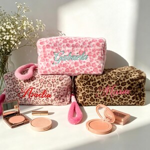 May include: A collection of personalized makeup bags in pink and brown leopard print. The bags are embroidered with names in various colors. Makeup items, including blush and lipstick, are arranged in front of the bags.