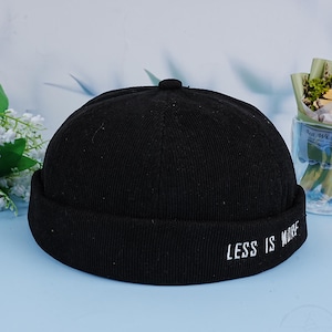 May include: Black ribbed knit beanie with a folded brim and the text "LESS IS MORE" embroidered in white on the brim.