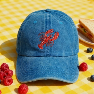 May include: A blue baseball cap with a red embroidered lobster design. The cap is made of a soft, faded fabric and has a curved brim. The cap is a casual accessory.