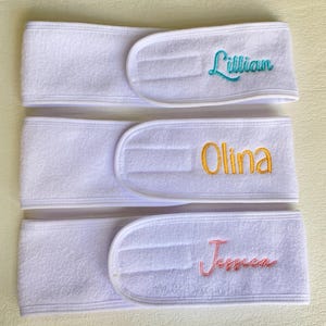 May include: Three white terry cloth headbands with embroidered names: "Lillian" in teal, "Olina" in gold, and "Jessica" in pink. The headbands have a hook and loop closure for adjustability. The text "Molly Designs Land" is printed at the bottom.