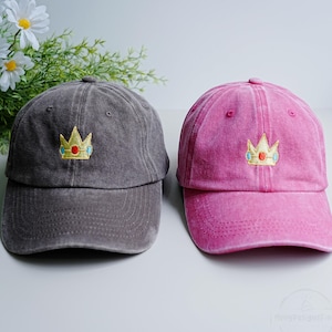 Puede incluir: Se muestran dos gorras de béisbol, una gris y otra rosa. Cada gorra presenta una corona dorada bordada con detalles azules y rojos. Las gorras están hechas de un material suave y texturizado, adecuado para uso casual.