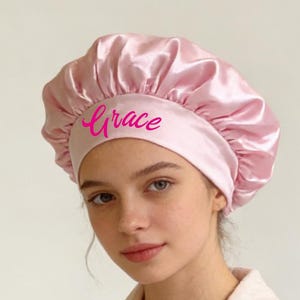 May include: A light pink satin bonnet with the name "Grace" embroidered in a matching pink script. The bonnet is worn with a light pink robe. The bonnet is designed to protect hair while sleeping.