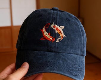 May include: A navy blue baseball cap featuring an embroidered design of two koi fish in shades of red, orange, and white. The cap has a curved brim and a small logo on the side.