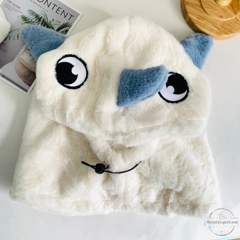 May include: White plush hooded hat with a blue and white cartoon rhino face. The hat has a black drawstring.