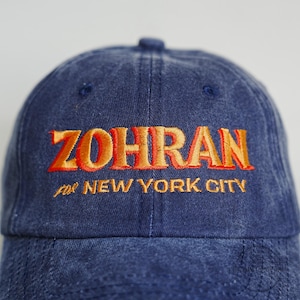 Pu&ograve; includere: Un cappellino da baseball in denim blu con il nome "ZOHRAN" ricamato con filo arancione. Sotto il nome, anche il testo "for NEW YORK CITY" &egrave; ricamato in arancione. Il cappellino ha una visiera curva e un bottone sulla parte superiore.