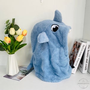 May include: A light blue plush hooded animal head shaped like a rhinoceros with a black eye and white teeth. The hood is made of soft, fuzzy fabric.