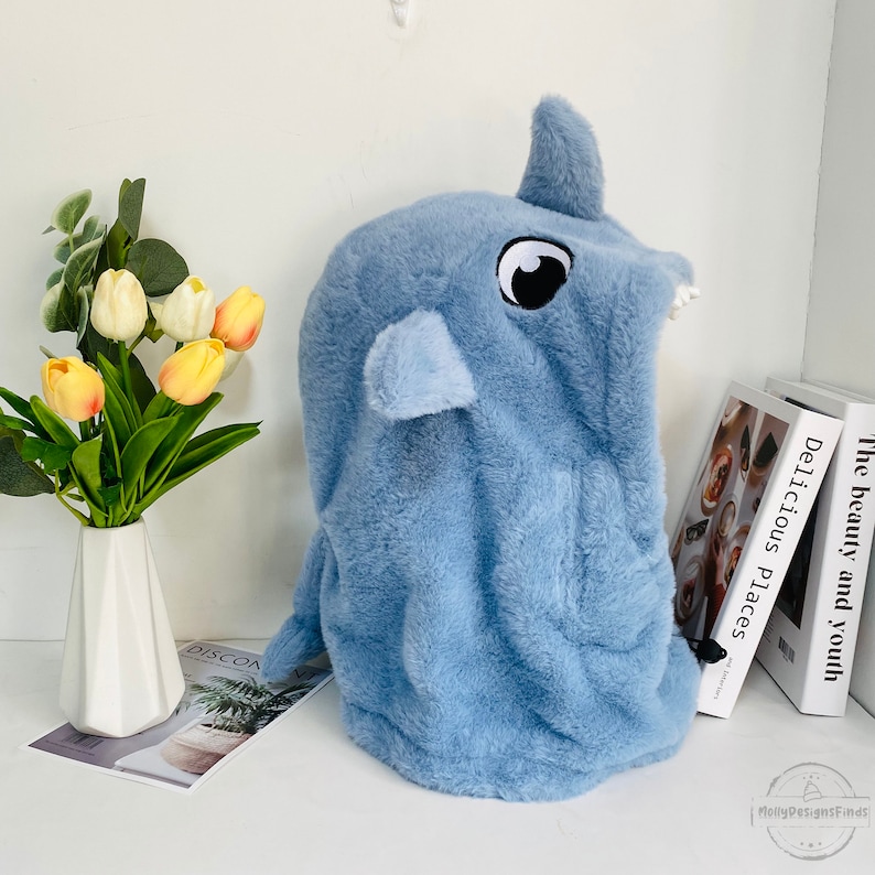 May include: A light blue plush hooded animal head shaped like a rhinoceros with a black eye and white teeth. The hood is made of soft, fuzzy fabric.