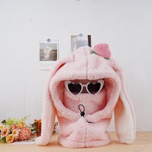 May include: Pink fuzzy bunny ear hooded hat with a peach on top and heart-shaped sunglasses. The hat has a drawstring closure.