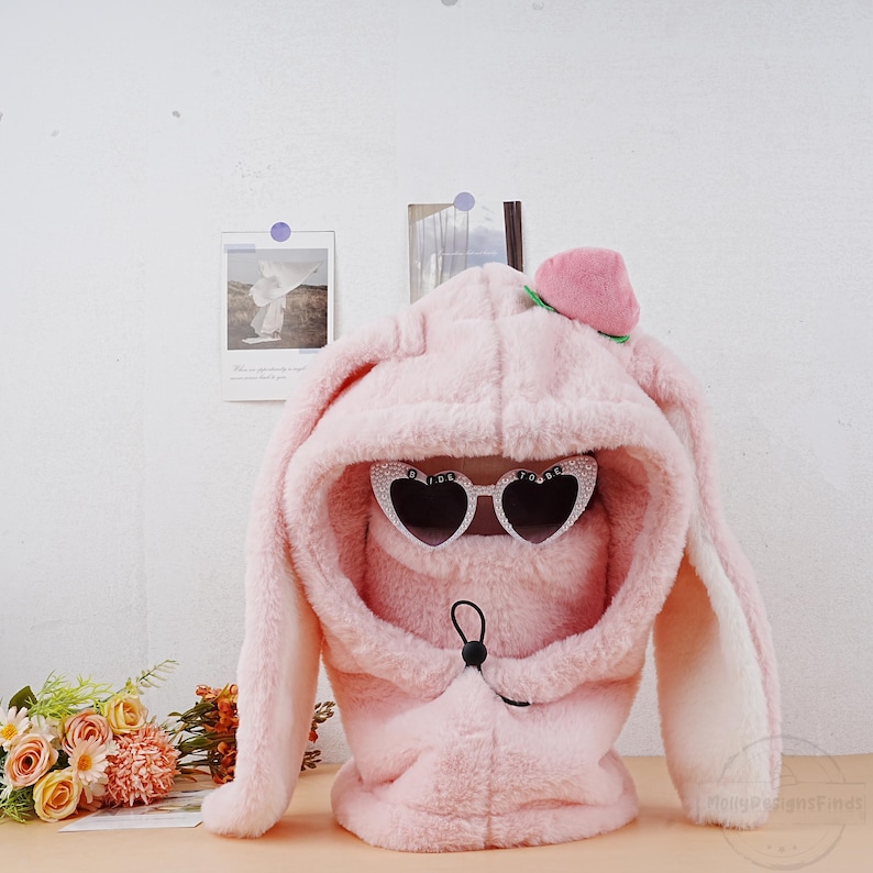 May include: Pink fuzzy bunny ear hooded hat with a peach on top and heart-shaped sunglasses. The hat has a drawstring closure.