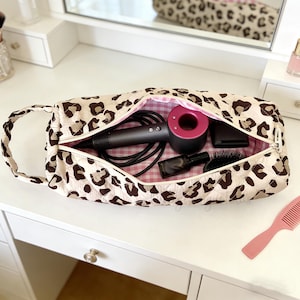 Könnte beinhalten: Eine Kosmetiktasche mit Leopardenmuster und rosa Vichy-Innenfutter, die einen schwarz-rosa Haartrockner, eine schwarze Bürste und andere Schönheitsaccessoires enthält. Die Tasche ist geöffnet und zeigt den Inhalt.