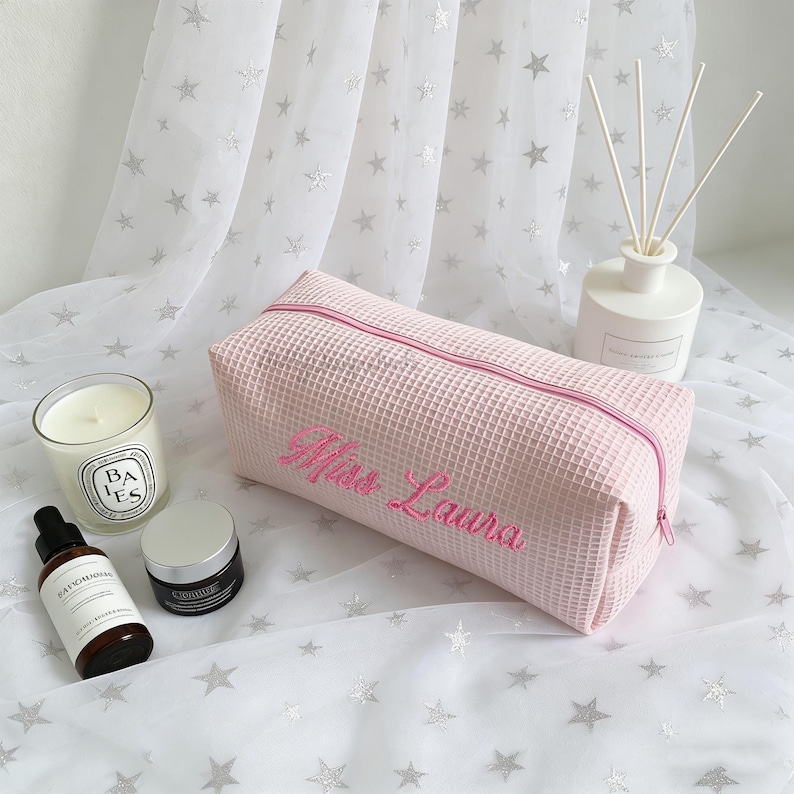 May include: A pink waffle-textured cosmetic bag with a matching zipper and the embroidered name "Miss Laura" in pink script. The bag is rectangular and sits on a white surface with a star pattern. Other items include a candle and skincare products.