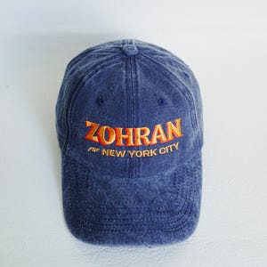 Pu&ograve; includere: Cappellino da baseball blu con la scritta ricamata "ZOHRAN for NEW YORK CITY" in arancione. Il cappellino ha una visiera curva e un bottone sulla parte superiore. Il tessuto ha un aspetto leggermente sbiadito.