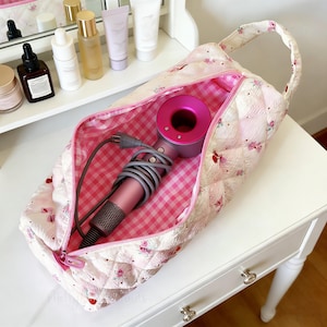 May include: A pink quilted cosmetic bag with a floral pattern, open to reveal a pink and gray hair dryer. The bag has a pink gingham lining and a zipper closure. The bag is on a white table.