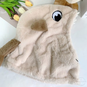 May include: A beige plush hooded animal costume with a brown tail and a black eye patch. The hood has a pointed snout and a small brown ear.