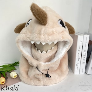 May include: A plush beige hooded hat shaped like a shark with a brown nose and black eyes. The hat has a white lining and a black drawstring.