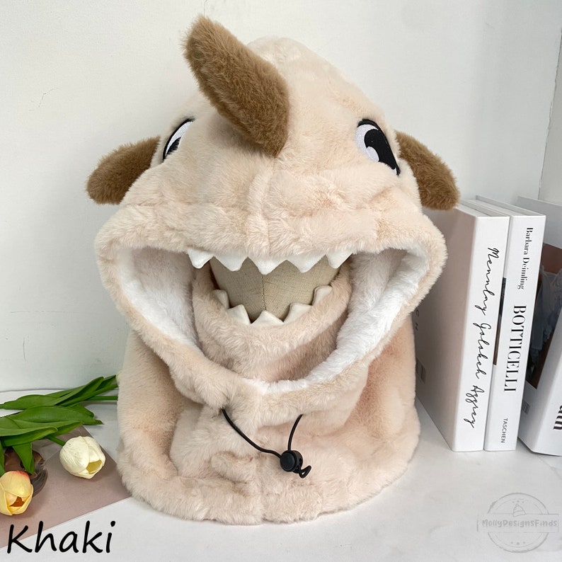 May include: A plush beige hooded hat shaped like a shark with a brown nose and black eyes. The hat has a white lining and a black drawstring.