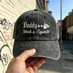 Könnte beinhalten: Eine anthrazitfarbene Baseballkappe in Acid-Wash-Optik mit weißem Sticktext Daddy Needs a Cigarette, ergänzt durch zwei rautenförmige Symbole und eine kleine Grafik einer Hand, die eine Zigarette hält, auf der Vorderseite.