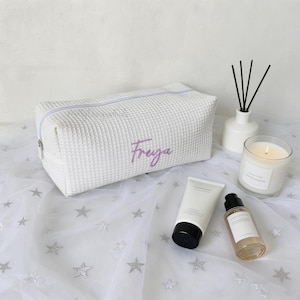 May include: White textured cosmetic bag with the name "Freya" embroidered in purple. The bag is rectangular with a zipper closure. A lit candle, diffuser, and skincare products are also visible.