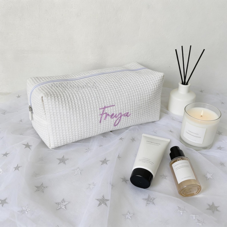 May include: White textured cosmetic bag with the name "Freya" embroidered in purple. The bag is rectangular with a zipper closure. A lit candle, diffuser, and skincare products are also visible.