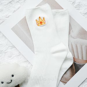 May include: A pair of white ribbed socks with a gold and pink embroidered crown design. The socks are made of a soft material and are suitable for everyday wear. The crown is a cute and whimsical detail.