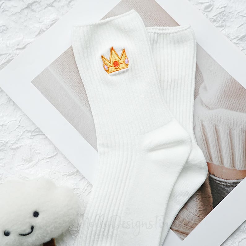 May include: A pair of white ribbed socks with a gold and pink embroidered crown design. The socks are made of a soft material and are suitable for everyday wear. The crown is a cute and whimsical detail.