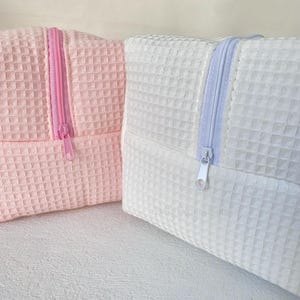 May include: Two small, square cosmetic bags. One is light pink with a pink zipper, and the other is white with a light blue zipper. Both bags have a waffle-weave texture.
