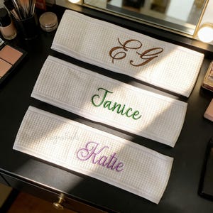 May include: Three white waffle-weave headbands with embroidered text. The top headband has brown initials, the middle one has the name "Janice" in green, and the bottom one has the name "Katie" in purple. The headbands are on a black surface.