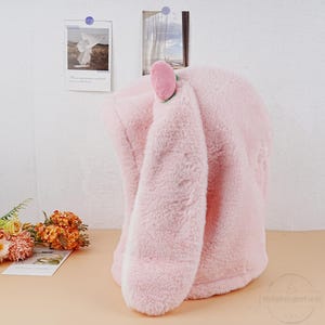 May include: Pink plush hooded jacket with long bunny ears. The ears are lined with white fur and have a pink felt flower on the top.