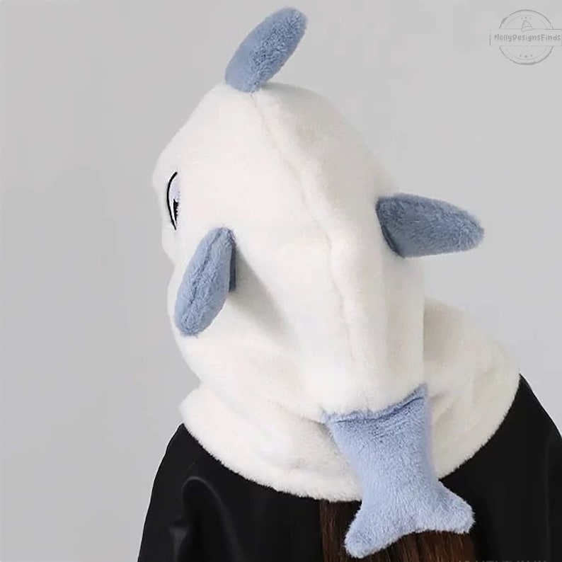 May include: White plush hooded hat with a blue dolphin design. The hood has a white plush body with blue plush fins and tail.