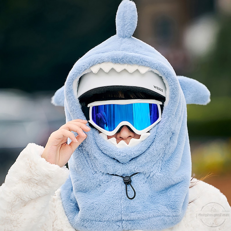May include: A light blue plush shark hooded face mask with a white helmet and blue ski goggles. The mask has a white drawstring.
