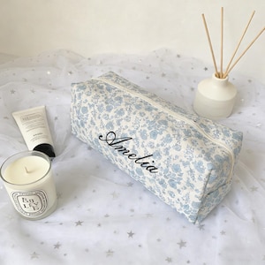 May include: A rectangular, floral-patterned cosmetic bag in light blue and white, with the name "Amelia" embroidered in black script. A white candle, a tube of lotion, and a white diffuser with reeds are also visible.