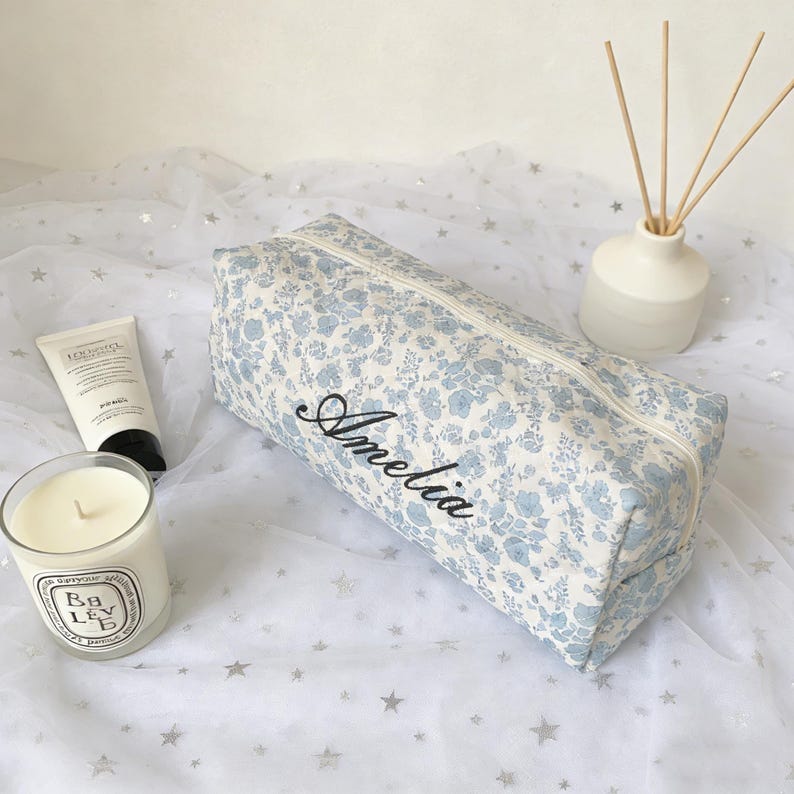 May include: A rectangular, floral-patterned cosmetic bag in light blue and white, with the name "Amelia" embroidered in black script. A white candle, a tube of lotion, and a white diffuser with reeds are also visible.