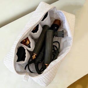 Peut inclure: Un sac à cosmétiques blanc texturé avec une fermeture éclair, ouvert pour révéler des outils de coiffure. Le sac contient une variété de bigoudis et d'outils de coiffure. Le sac est en tissu gaufré.