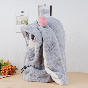 May include: A gray plush bunny-shaped hooded blanket with white trim and a pink peach on top. The hood has a pair of white sunglasses.
