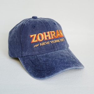 Pu&ograve; includere: Un cappellino da baseball in denim blu con la scritta ricamata "ZOHRAN" in arancione e giallo, con "FOR NEW YORK CITY" sotto. Il cappellino ha una visiera curva e un bottone sulla parte superiore.