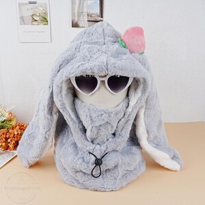May include: Gray plush bunny hood with long ears, a white faux fur trim, and a pink peach flower accent. The hood has a drawstring closure and features heart-shaped sunglasses.