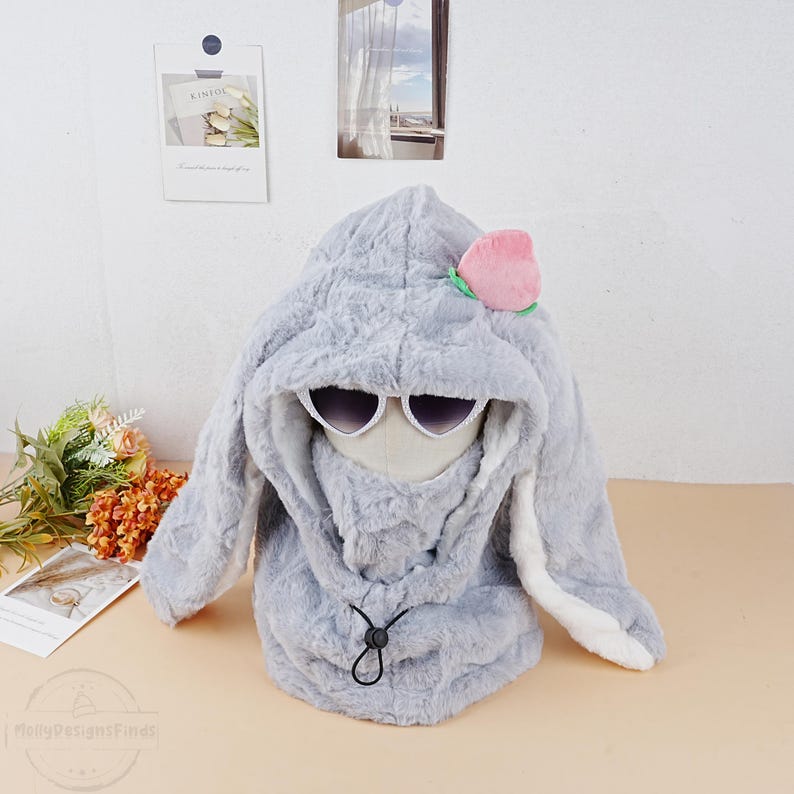 May include: Gray plush bunny hood with a pink peach flower accent and white trim on the ears. The hood has a black drawstring and a pair of sunglasses.