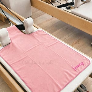 May include: A rectangular pink pilates reformer towel with rounded top corners and a matching pink stitched border. The towel features the embroidered name Lainey in a cursive script in the bottom right corner and the text MollyDesignsfinds.