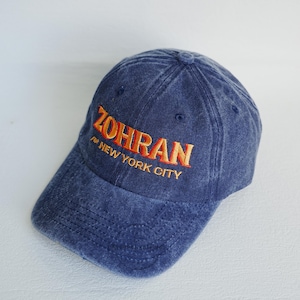Pu&ograve; includere: Un cappellino da baseball in denim blu con la parola "ZOHRAN" ricamata in filo arancione. Sotto la parola "ZOHRAN" c'&egrave; la frase "for NEW YORK CITY". Il cappellino ha una visiera curva e un design a sei pannelli.