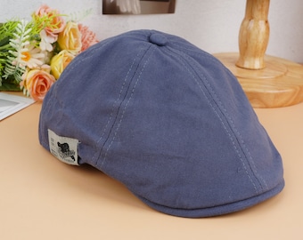 May include: A blue newsboy cap with a rounded crown and a small brim. The cap has a stitched design and a small tag with text and a bear graphic. The cap is made of a soft fabric.