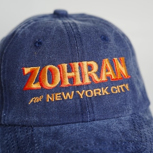 Pu&ograve; includere: Un cappellino da baseball blu navy con la scritta "ZOHRAN" ricamata in filo arancione e rosso. Sotto, la frase "FOR NEW YORK CITY" &egrave; anch'essa ricamata in arancione. Il cappellino &egrave; realizzato in un tessuto morbido e usurato.