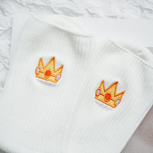 May include: A pair of white ribbed socks with embroidered gold crowns featuring orange and purple accents. The socks are made of a soft, comfortable material and are suitable for everyday wear.