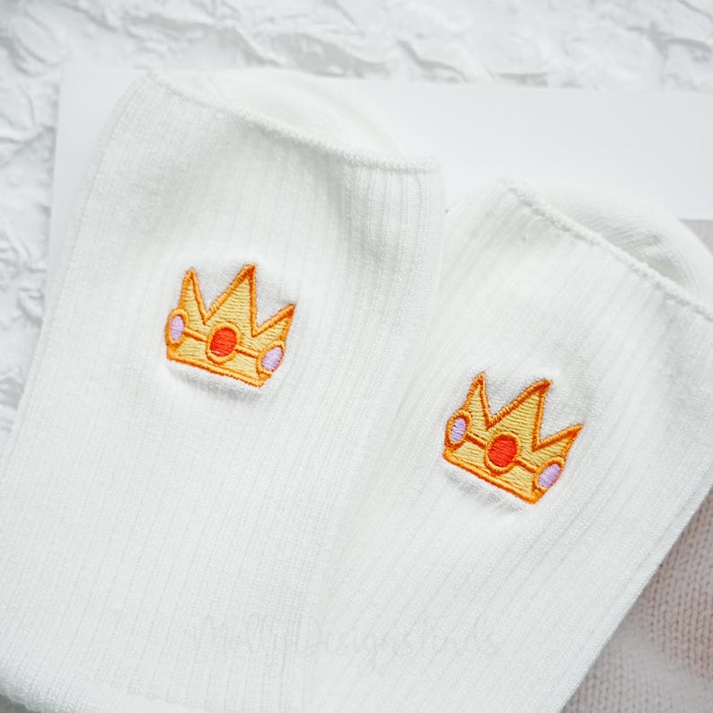 May include: A pair of white ribbed socks with embroidered gold crowns featuring orange and purple accents. The socks are made of a soft, comfortable material and are suitable for everyday wear.