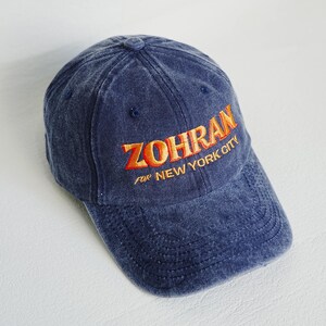 Pu&ograve; includere: Un cappellino da baseball blu navy con la scritta ricamata "ZOHRAN for NEW YORK CITY" in arancione. Il cappellino ha una visiera curva e un aspetto leggermente sbiadito.