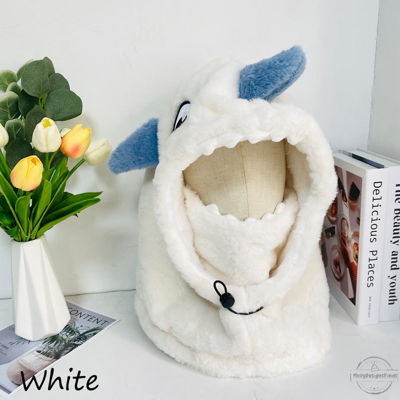 May include: White plush hooded scarf with a blue and white monster face design. The scarf has a drawstring closure and is made of soft, fluffy material.