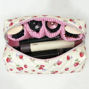 Puede incluir: Un neceser acolchado con estampado de fresas, abierto para revelar herramientas y cepillos de peinado. El neceser tiene un interior de cuadros rosas y un cierre de cremallera. El exterior es de color crema con fresas rojas y hojas verdes.
