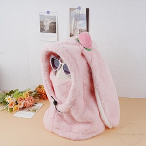 May include: Pink plush bunny hood with white trim and a peach-shaped detail on top. The hood has a drawstring and a pair of heart-shaped sunglasses are peeking out from the hood.